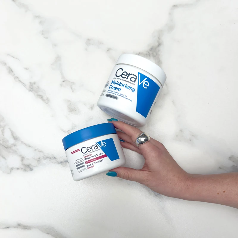 A hand with teal nails holds two CeraVe moisturising cream jars on a marble surface. Both jars are white with blue labels.