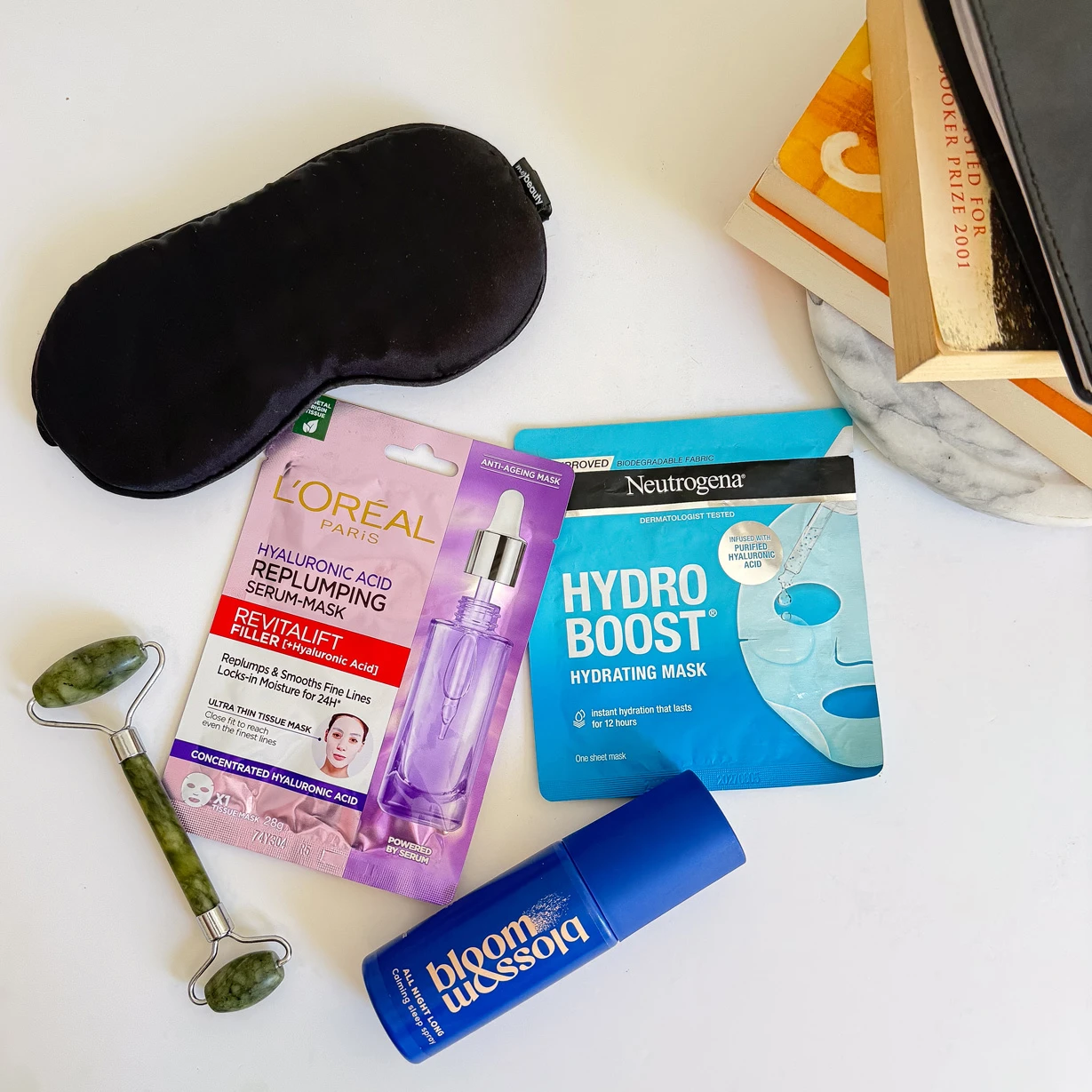 Skincare products including face masks, jade roller, and sleep mask arranged on white surface with beauty book in background