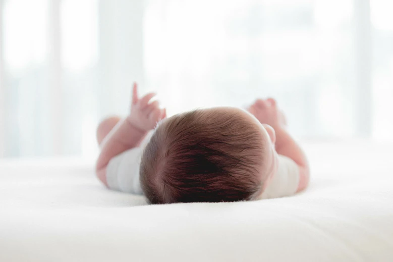 Newborn baby lying on white bedding, viewed from behind showing dark hair and tiny raised feet in soft lighting