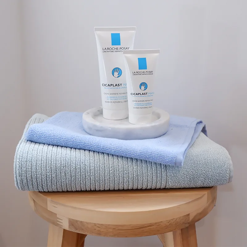 Two La Roche-Posay Cicaplast tubes on marble tray, displayed atop folded blue and gray towels on wooden stool.