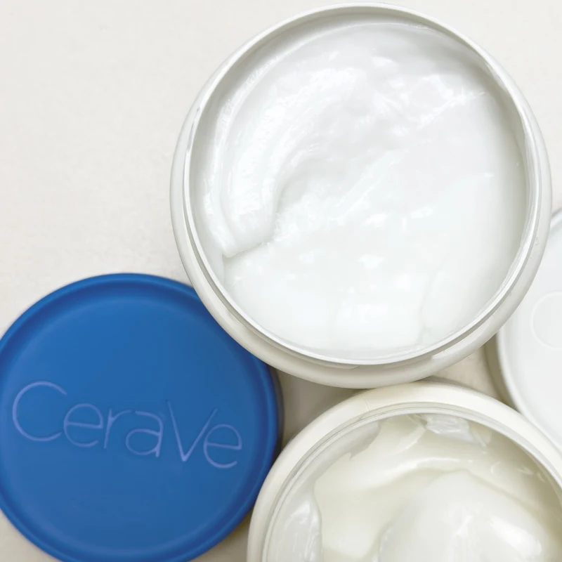 Two open CeraVe moisturizing cream jars showing white cream inside, with a blue CeraVe lid placed beside them.