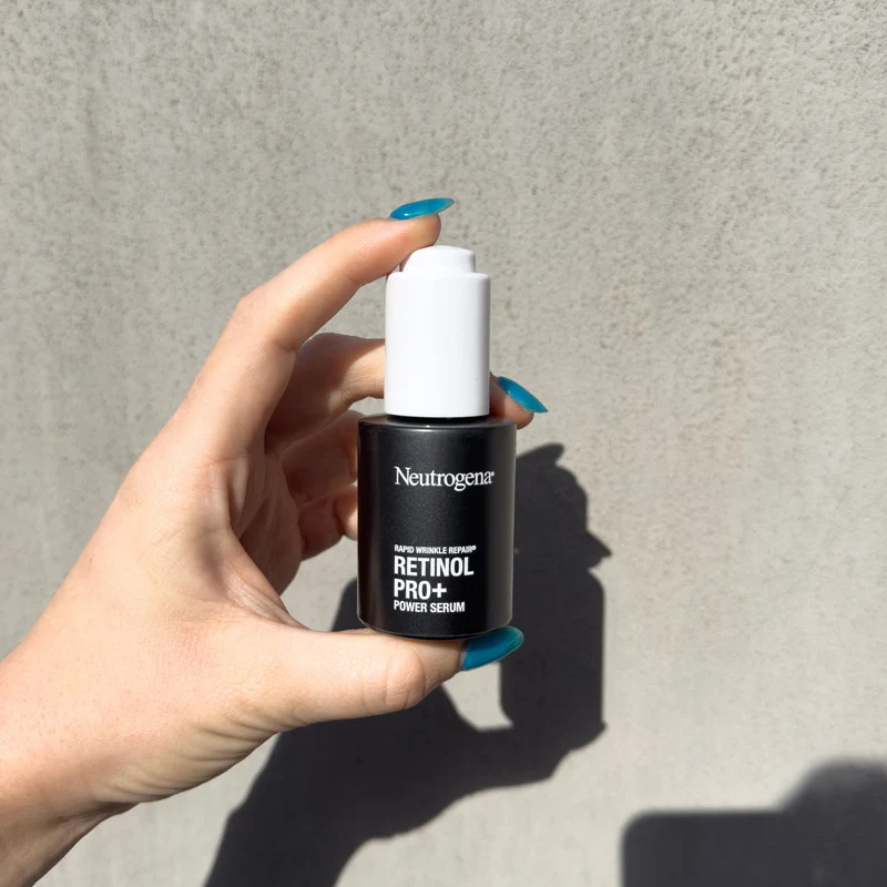 Hand with teal nails holding a dark bottle of Neutrogena Rapid Wrinkle Repair Retinol Pro+ Power Serum against a gray wall.