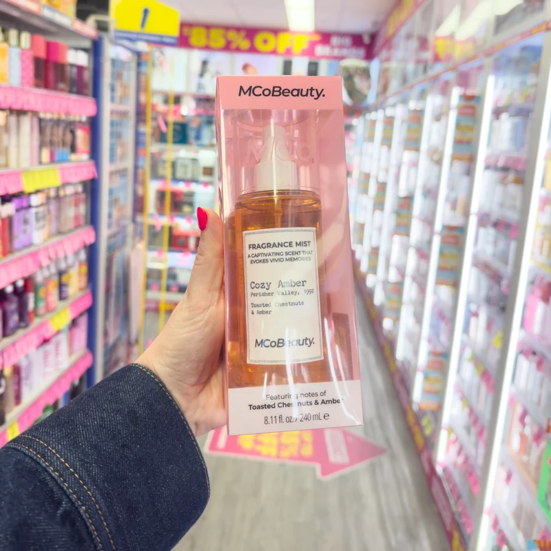 Hand holding MCoBeauty Cozy Amber Fragrance Mist in a pink box in a beauty store aisle with colorful shelves.