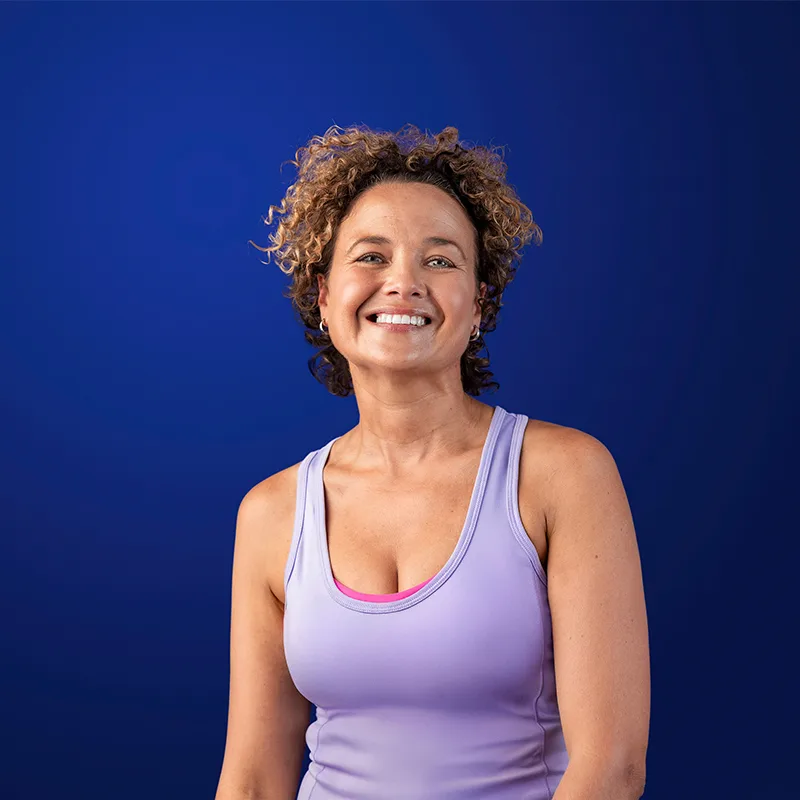 A smiling person with curly hair wearing a lavender athletic tank top against a deep blue background.
