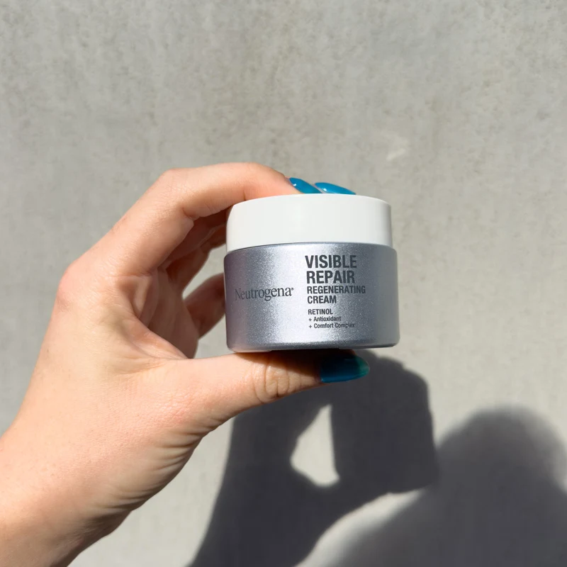 A hand with blue nails holds a Neutrogena Visible Repair Regenerating Cream jar against a light gray wall in sunlight.