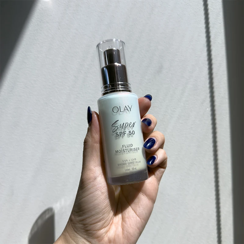 Hand with dark blue nail polish holding Olay Super SPF 50 fluid moisturizer bottle against white background with shadow lines.