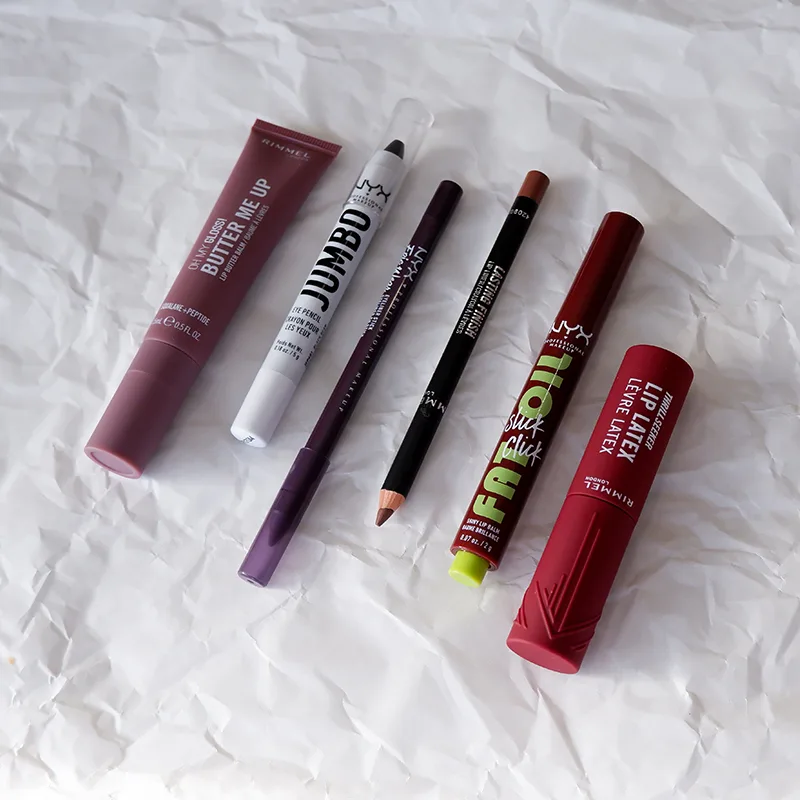 Various lip makeup products including Rimmel and NYX brand items arranged on crumpled white paper background. 