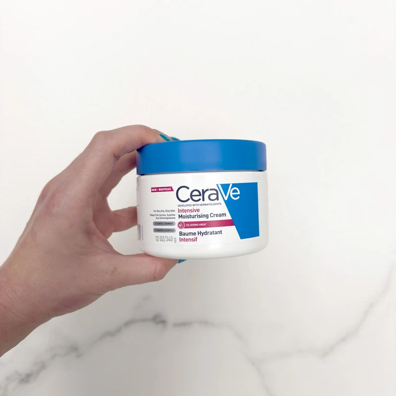 A hand holding a CeraVe Intensive Moisturising Cream jar with a blue lid, 12 oz, against a white marble background.