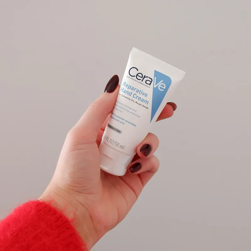 Hand with dark burgundy nails holding CeraVe Reparative Hand Cream tube against light gray background, wearing red sleeve.