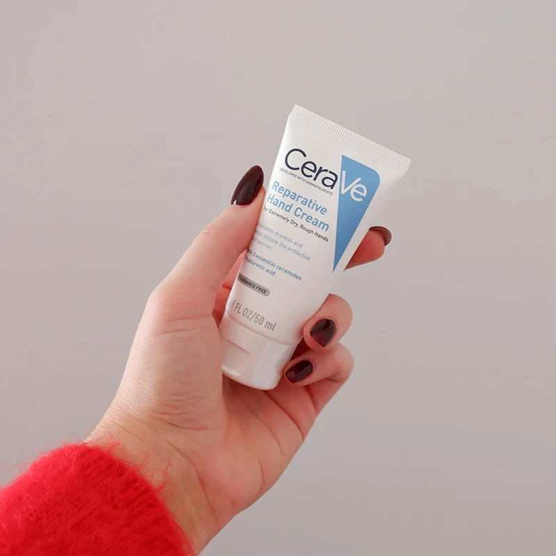 Hand with dark burgundy nails holding CeraVe Reparative Hand Cream tube against light gray background, wearing red sleeve.