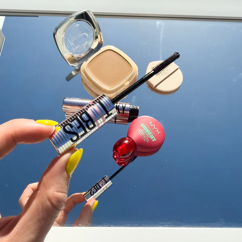 Hands with yellow nails hold a mascara wand. Nearby are a L'Oreal powder compact, NYX pink blush, and a red lip product.