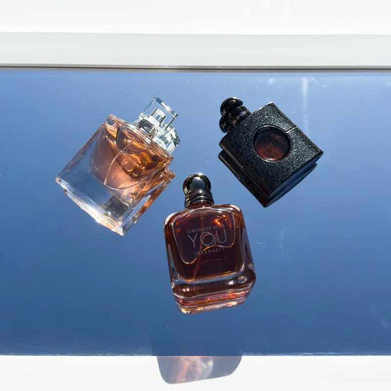 Three perfume bottles on a reflective blue surface: an amber glass bottle, a black textured bottle, and Armani Stronger With You Intensely.
