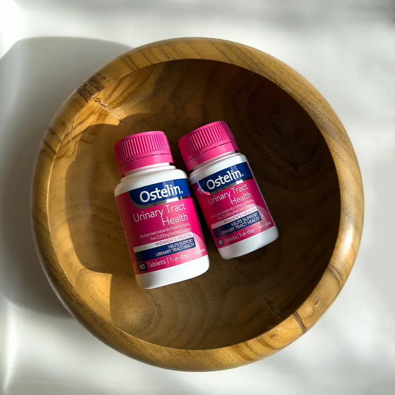 Two bottles of Ostelin Urinary Tract Health supplements with pink caps placed in a wooden bowl.