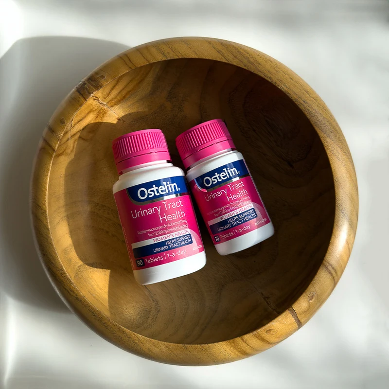 Two bottles of Ostelin Urinary Tract Health supplements with pink caps placed in a wooden bowl.