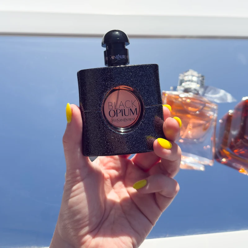 A hand with yellow nails holds a black glittery Yves Saint Laurent Black Opium perfume bottle against a bright blue background.