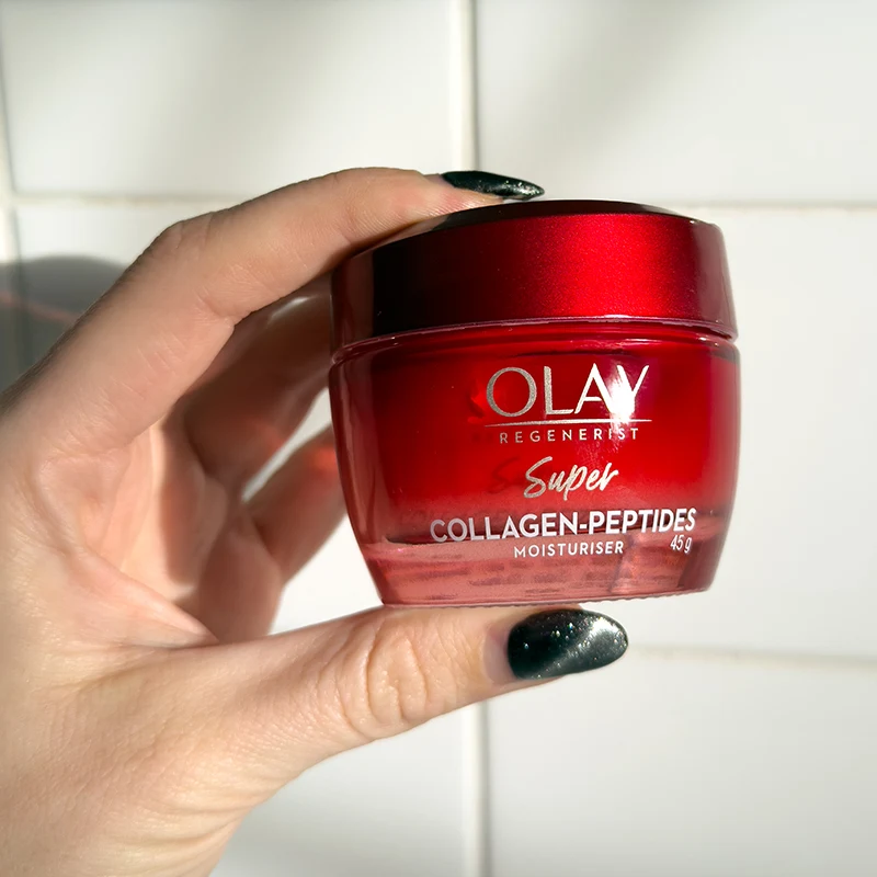 Hand with black nail polish holding a red jar of Olay Regenerist Super Collagen-Peptides moisturizer against white background.