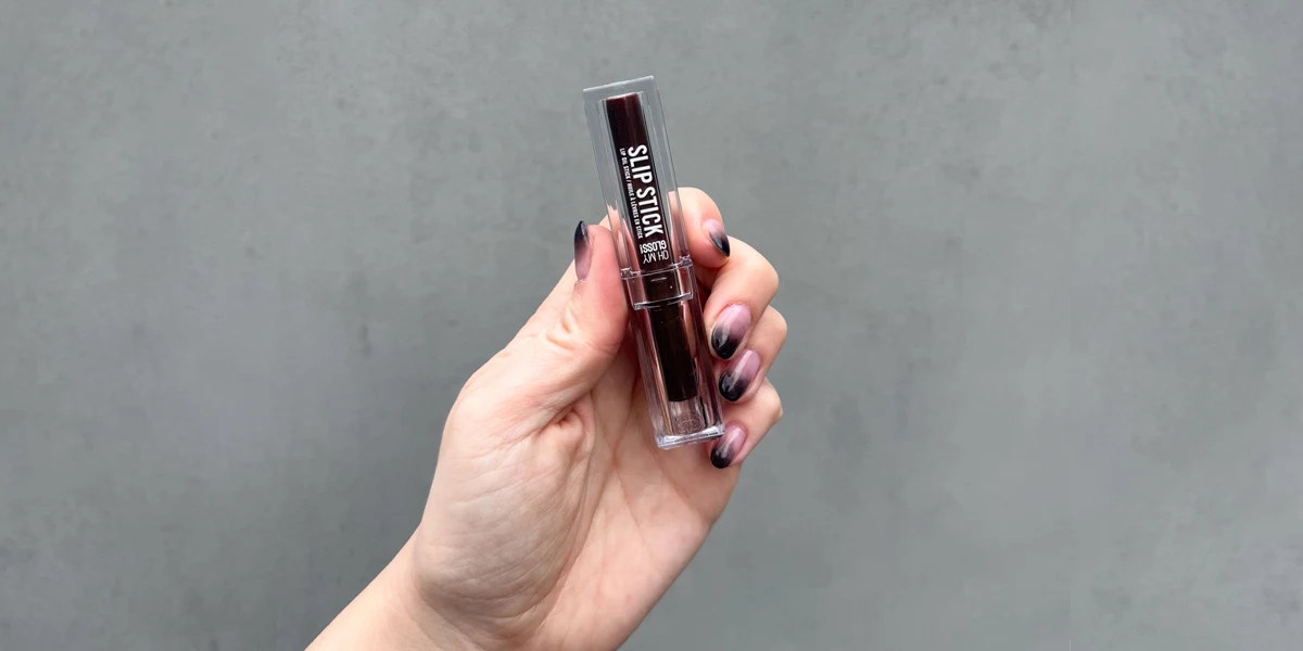 Hand with black ombre nails holding an Oh My Gloss Slip Stick lip product in a clear case against a gray background.