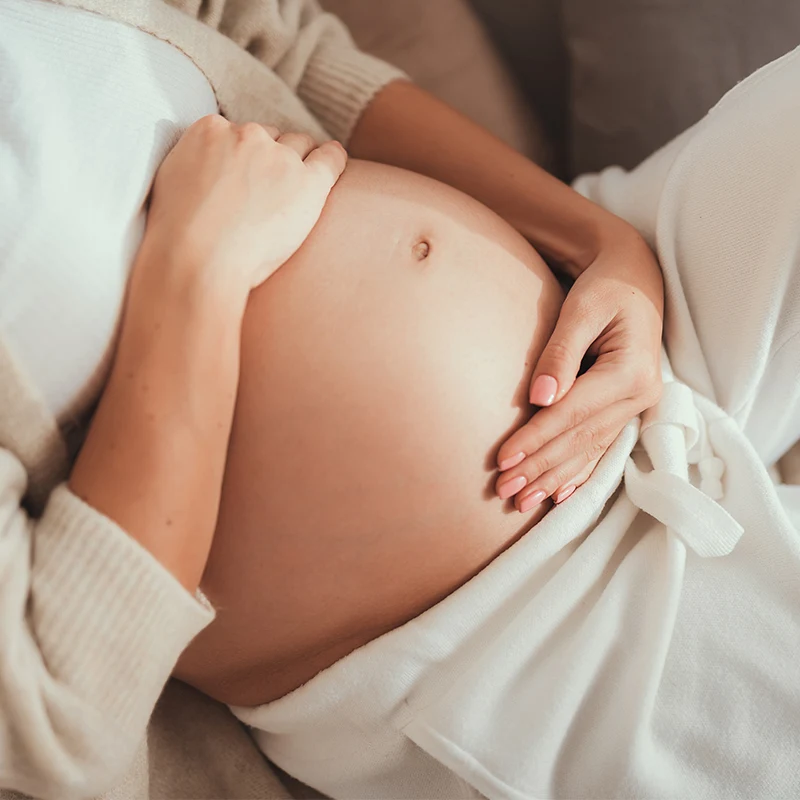 Close-up of pregnant belly with hands gently resting on it, wrapped in soft white fabric against light-colored bedding.