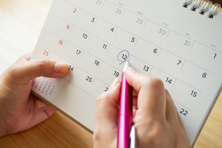 Hand holding pink pen circles date on spiral-bound monthly calendar, marking appointment or deadline