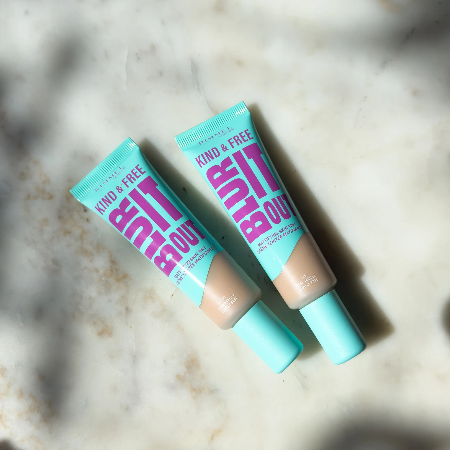 Two turquoise cosmetic tubes with pink text lying on a white marble surface, casting diagonal shadows
