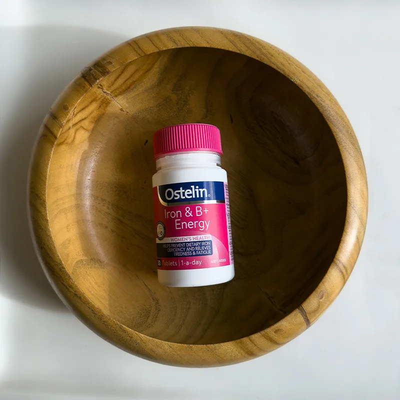 Bottle of Ostelin Iron & B+ Energy women's health supplements in a wooden bowl against white background.