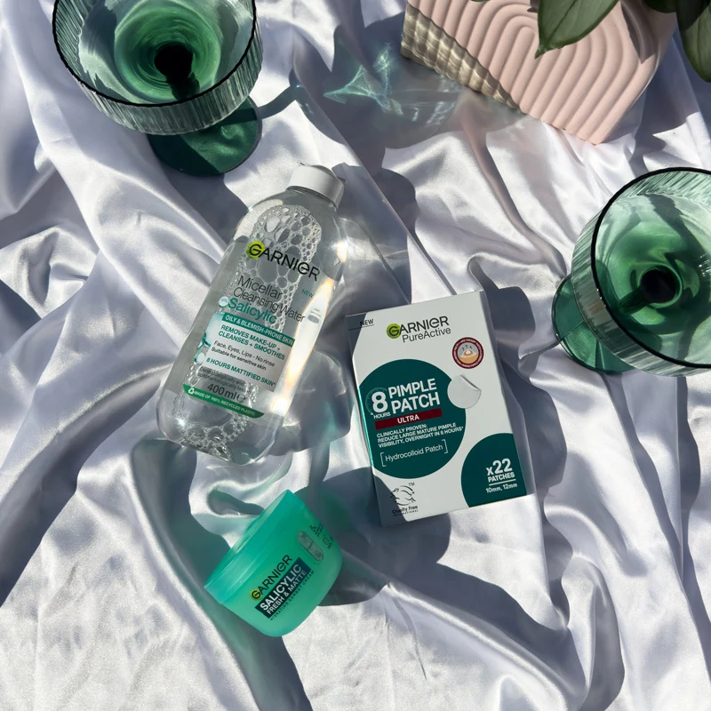 Three Garnier skincare products on white fabric: Micellar Cleansing Water, PureActive Pimple Patches, and a green Salicylic moisturiser.