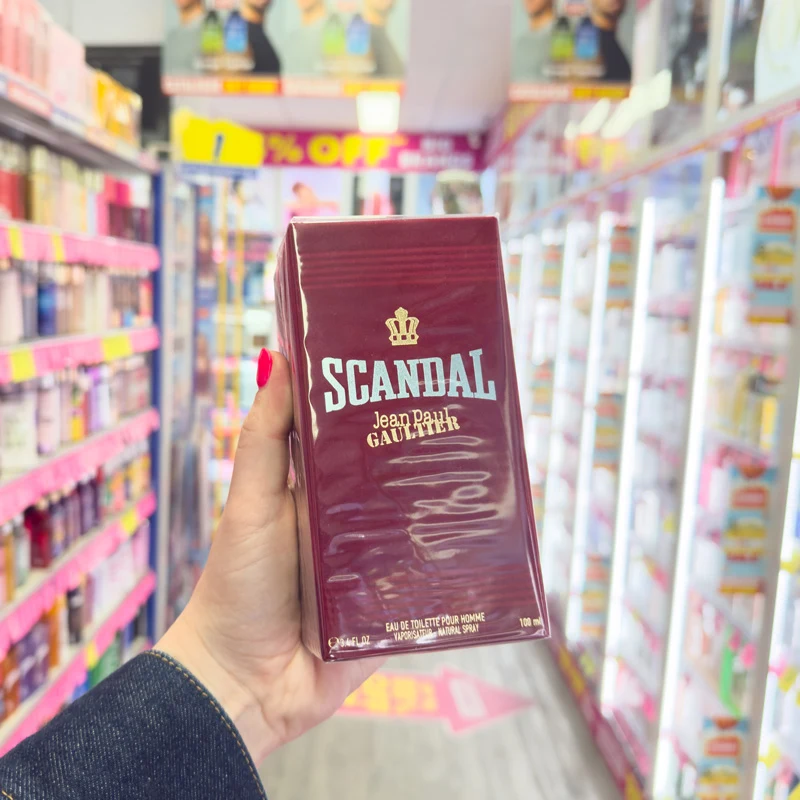 Hand holding Jean Paul Gaultier Scandal Pour Homme EDT 100ml in a dark red box inside a retail store aisle.
