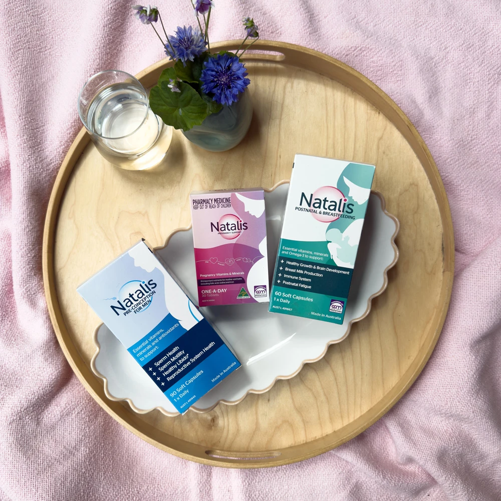 Three Natalis vitamin supplement boxes on wooden tray with blue cornflowers and glass of water, on pink fabric background