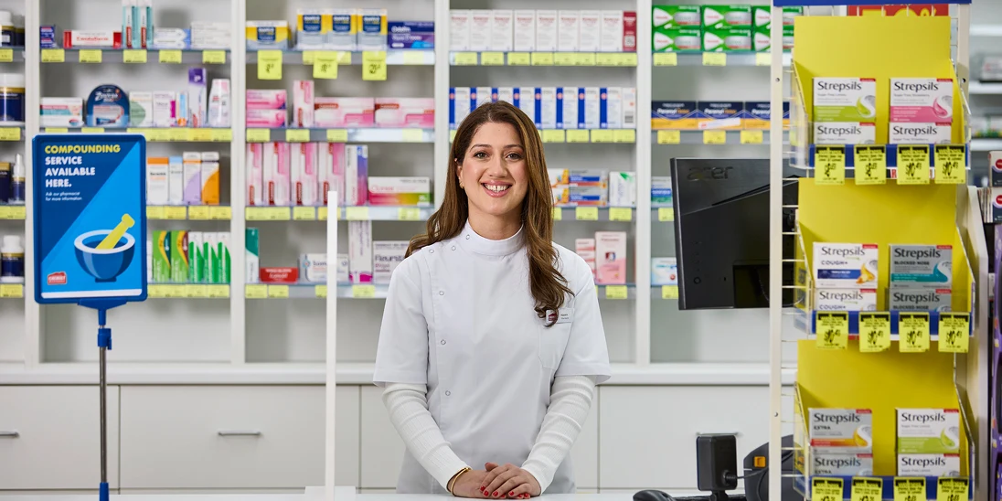 A smiling female pharmacist in a white coat stands at a pharmacy counter, surrounded by shelves of medications and Strepsils products.