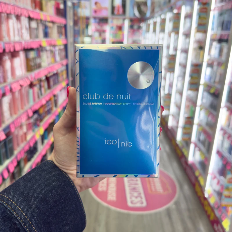 A hand holding a blue Club de Nuit Iconic Eau de Parfum box in a store aisle lined with colorful products.