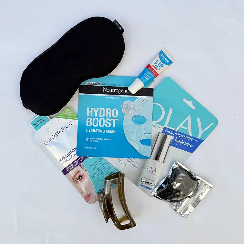 Travel skincare essentials laid flat: Embryolisse cream, Neutrogena hydrating mask, lip balm, and black sleep mask on white background.
