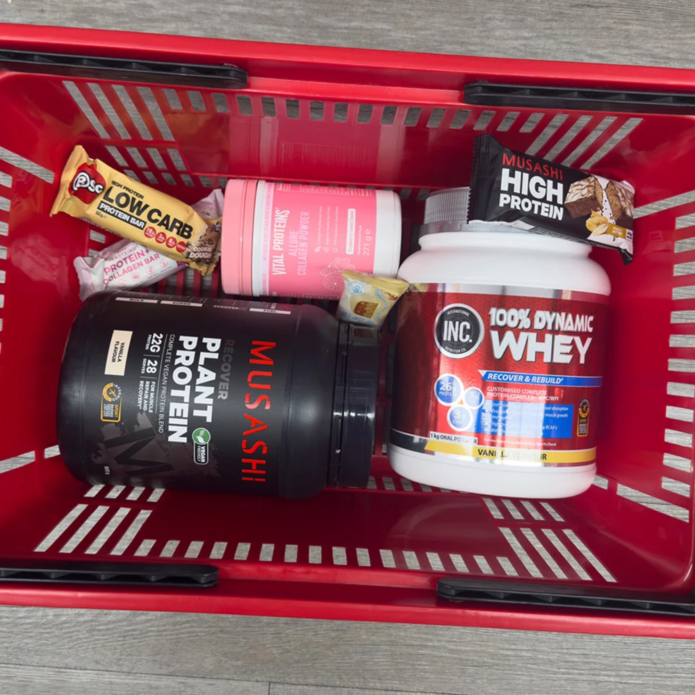 Red shopping basket filled with protein supplements including Musashi Plant Protein, INC Whey, Vital Proteins collagen, and protein bars.