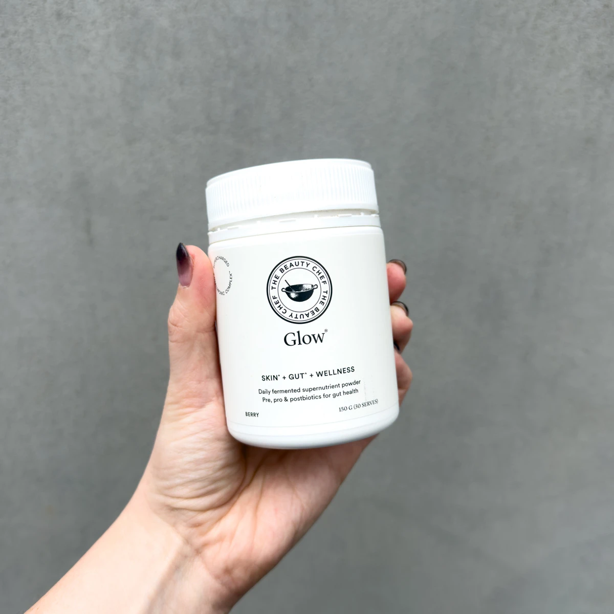 A hand holding a white tub of The Beauty Chef Glow berry-flavoured fermented supernutrient powder against a grey background.