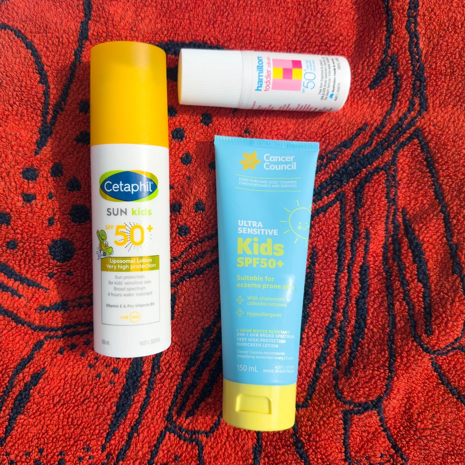 Three kids SPF50+ sunscreen products on a red towel: Cetaphil Sun Kids spray, Cancer Council Ultra Sensitive tube, and Hamilton Toddler roll-on.