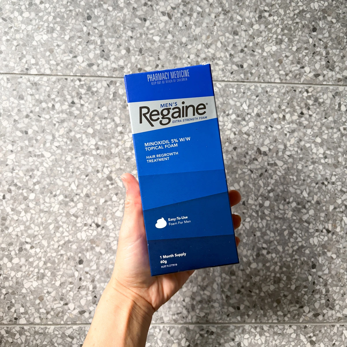 A hand holding a blue box of Men's Regaine Extra Strength Foam, a minoxidil 5% hair regrowth treatment, against a grey terrazzo background.