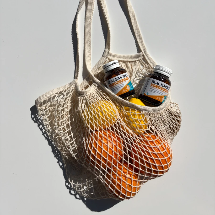 A cream mesh bag holding oranges, a lemon, and two Blackmores Gut Immune Biotic supplement bottles on a light grey background.