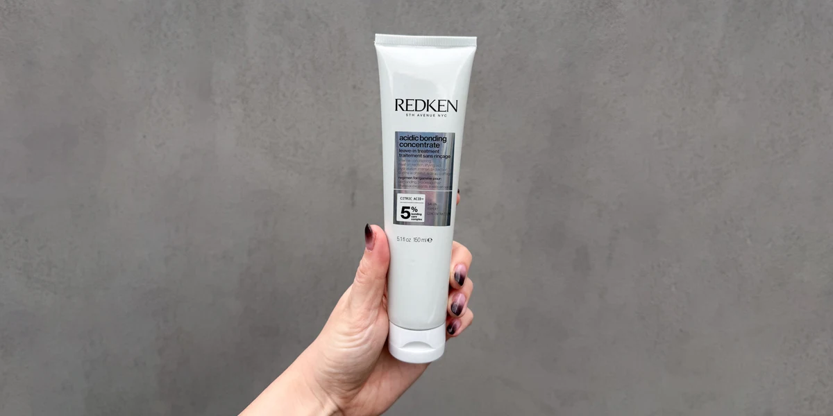 A hand holding a white tube of Redken Acidic Bonding Concentrate leave-in treatment, 150ml, against a gray background.