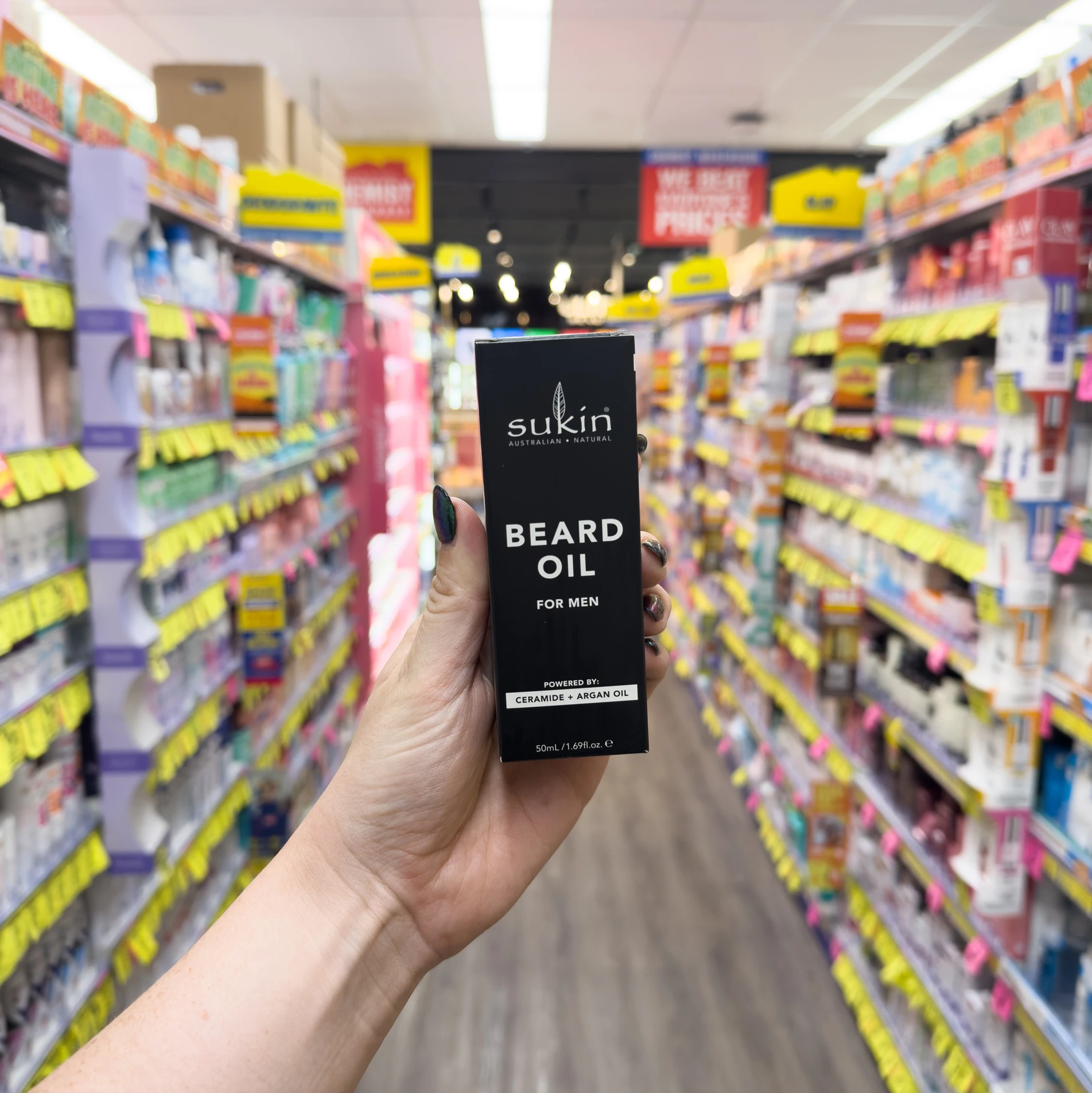 Hand holding Sukin beard oil product in black packaging against blurred retail store shelves with colorful product displays