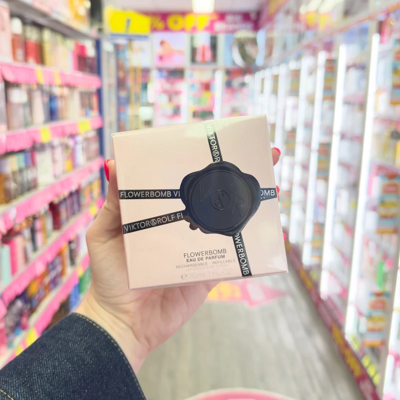 Hand holding Viktor&Rolf Flowerbomb Eau de Parfum box in a colorful retail store aisle with shelves in background.