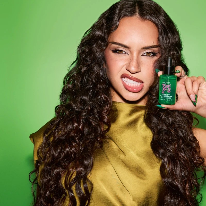 Katseye Daniella with long curly dark hair holds Matrix Food for Soft hair oil, wearing gold top, making a playful face on green background.