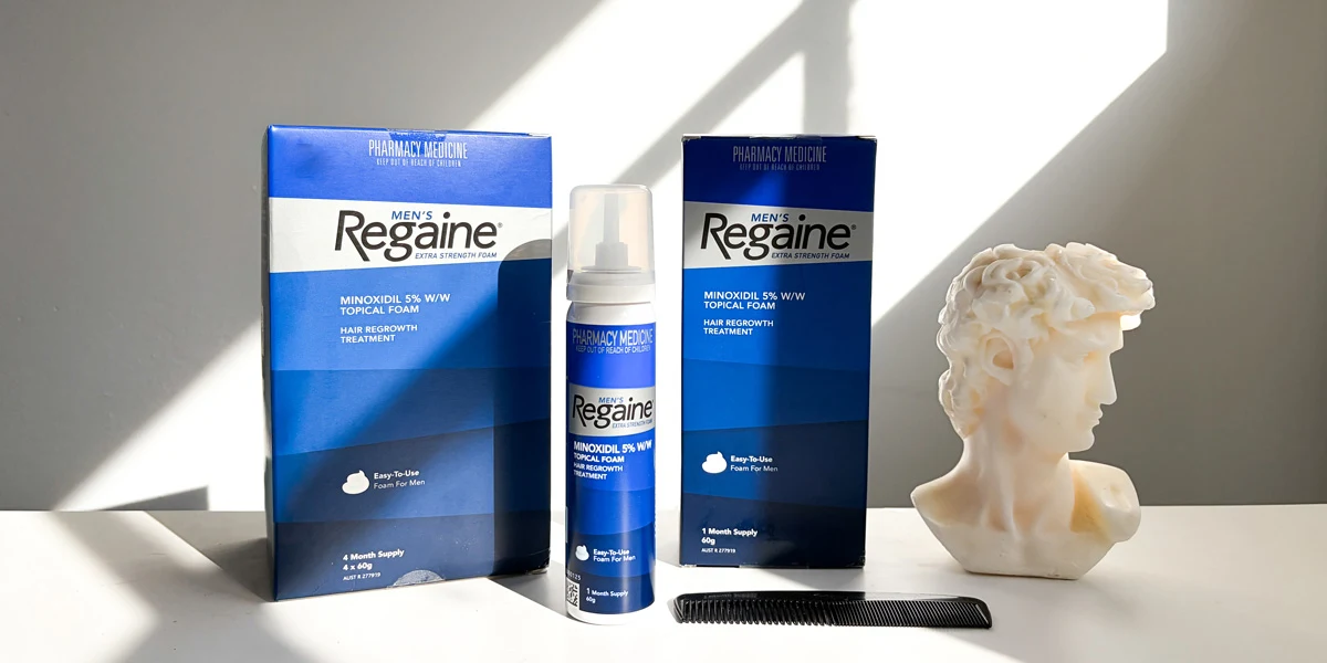 Two Men's Regaine hair regrowth foam boxes, a spray bottle, a black comb, and a white marble bust on a white surface.