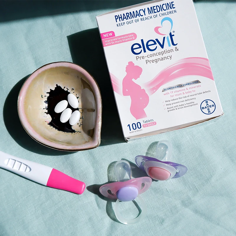 Elevit pregnancy vitamins box, heart-shaped dish with pills, pregnancy test, and baby pacifier arranged on light blue fabric.