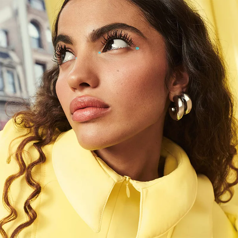 Close-up beauty portrait of woman with curly hair wearing yellow jacket, gold hoop earrings, and turquoise eye accent.
