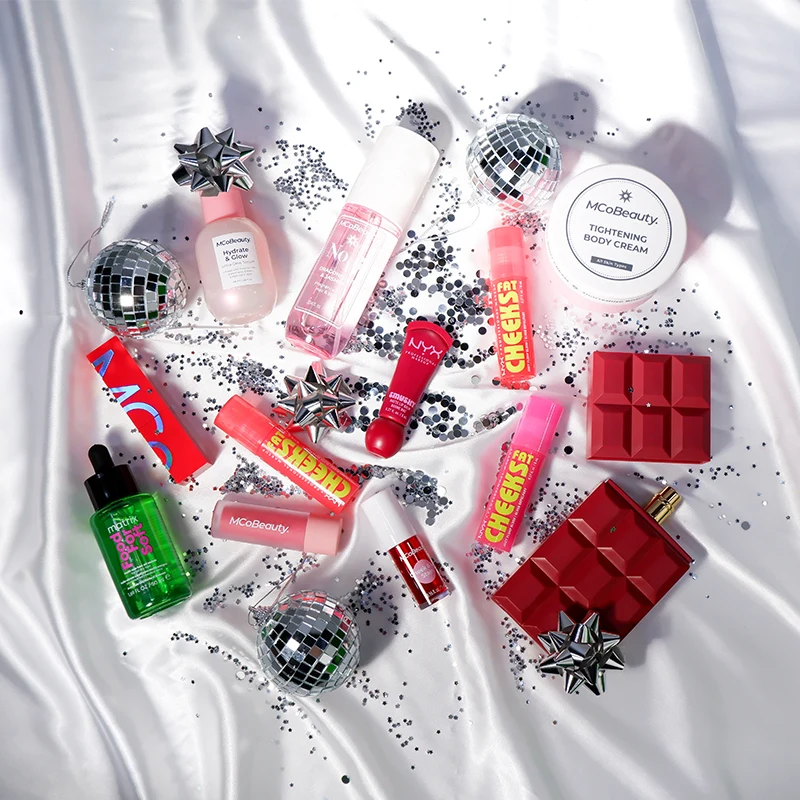 Beauty products and cosmetics arranged with disco balls, silver bows, and glitter on white satin, featuring pink and red holiday packaging.