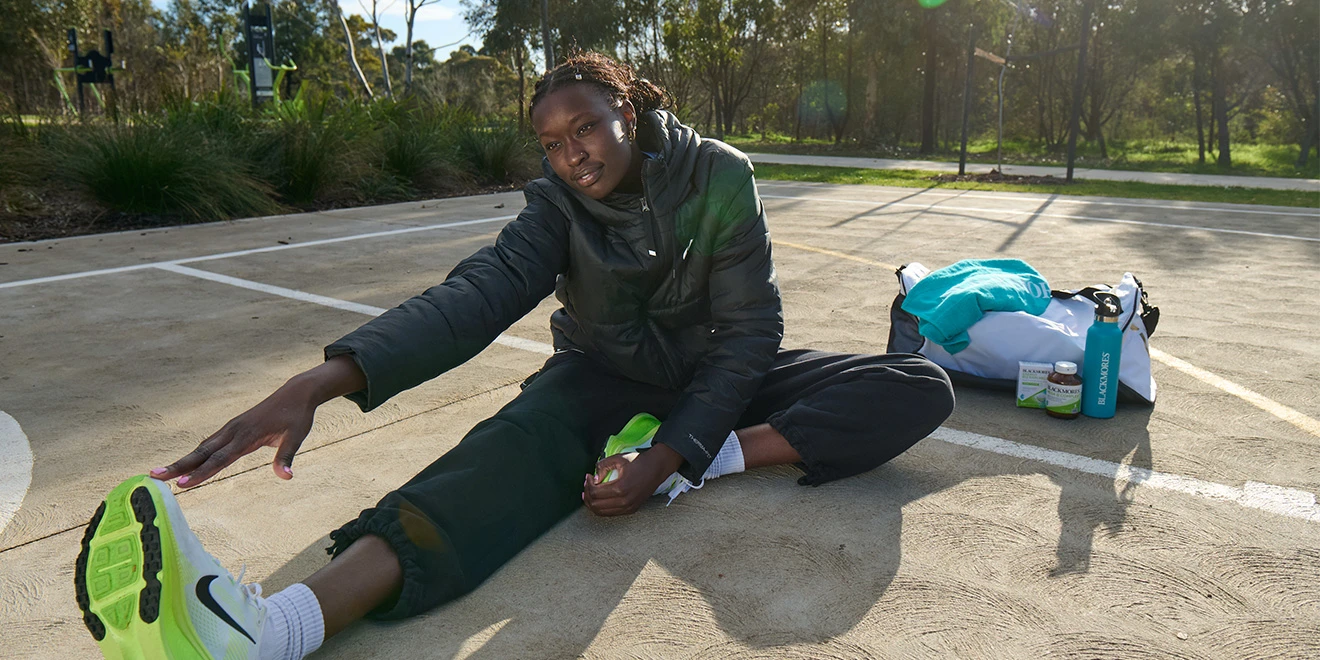 Student in black athletic wear stretches on concrete ground in sunny outdoor setting, reaching for toes with bright green sneakers.