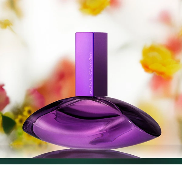 Calvin Klein Euphoria perfume bottle in purple, surrounded by orchids, vanilla sticks, and wood on a purple background.