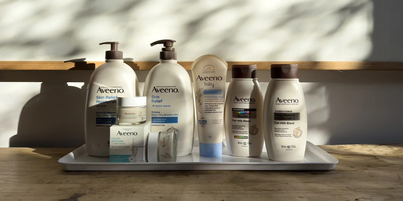 Collection of Aveeno skincare products arranged on wooden shelves, including body wash, moisturisers, and hair care items in white bottles.