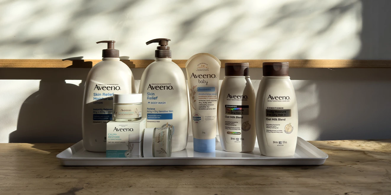 Collection of Aveeno skincare products arranged on wooden shelves, including body wash, moisturisers, and hair care items in white bottles.