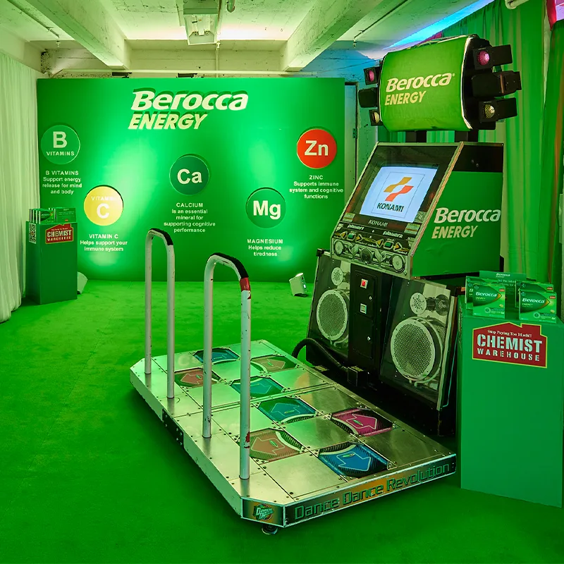 Dance Revolution arcade game setup in green-lit Berocca Energy promotional display with vitamin benefits shown on wall backdrop.