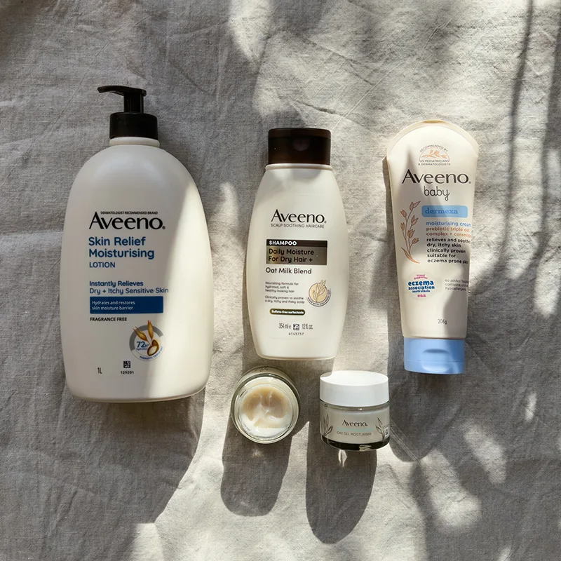 Collection of Aveeno skincare products including moisturising lotion, shampoo, baby cream, and face moisturisers on gray fabric.